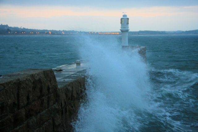 Waves breaking over the harbour arm beside the lighthouse at Penzance in Cornwall at dusk