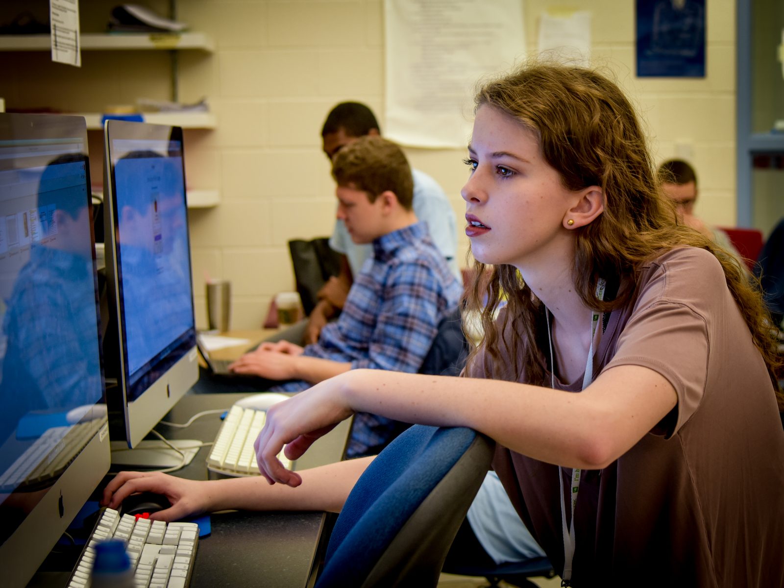 A student working at a desktop computer in a school classroom, with classmates at computers behind her