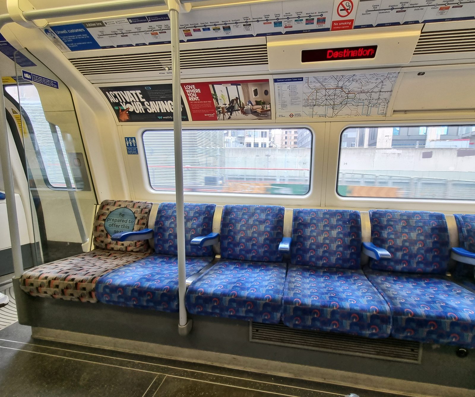 A row of empty blue patterned seats on a London Underground Jubilee line train, beneath line maps and a Tube map